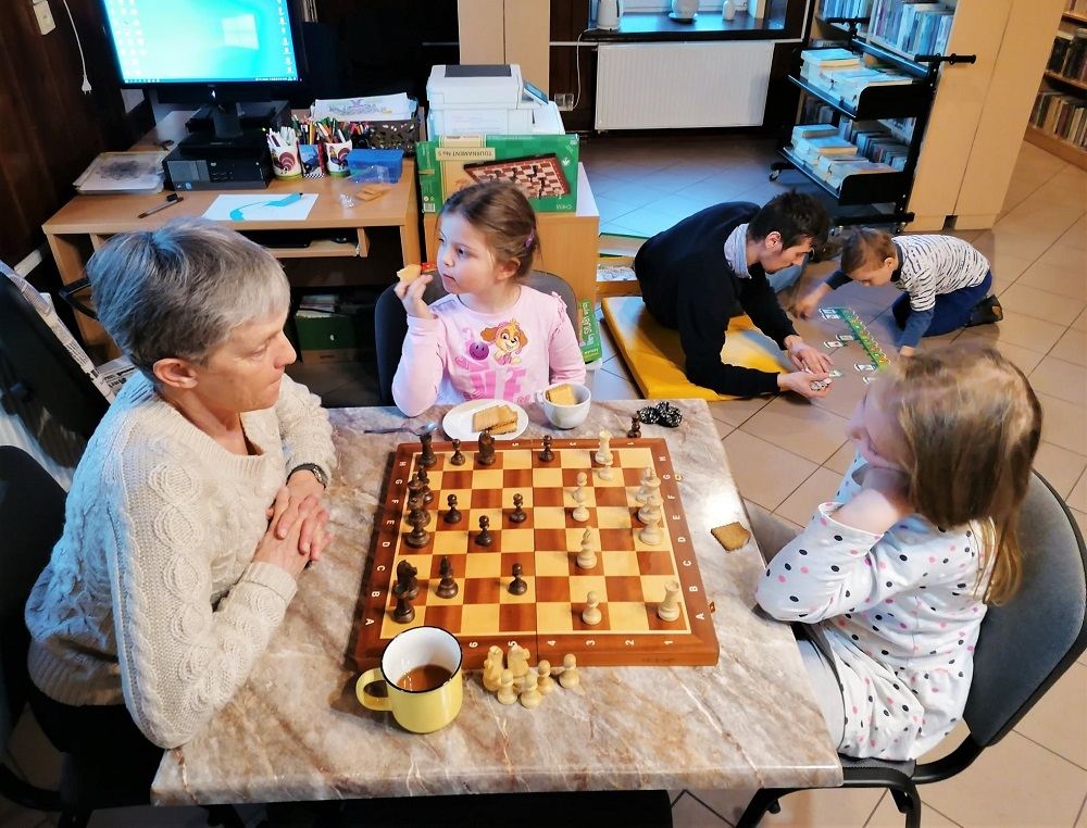 Instruktorka uczy dzieci gry w szachy. W tle zabawy rodzica z dzieckiem.