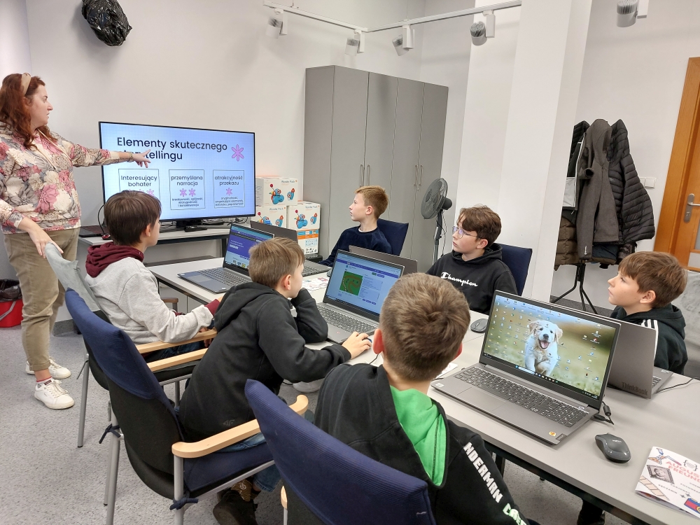 Grupa dzieci siedząca przy komputerach z uwagą słucha prowadzącej zajęcia bibliotekarki