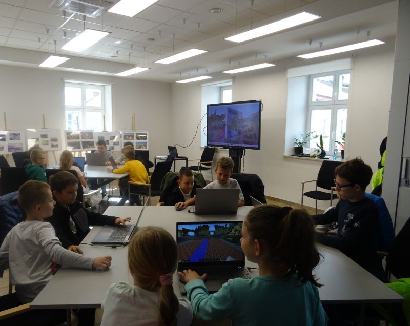 Dzieci siedzą przy stolikach na których rozłożone są komputery i grają w gre Minecraft