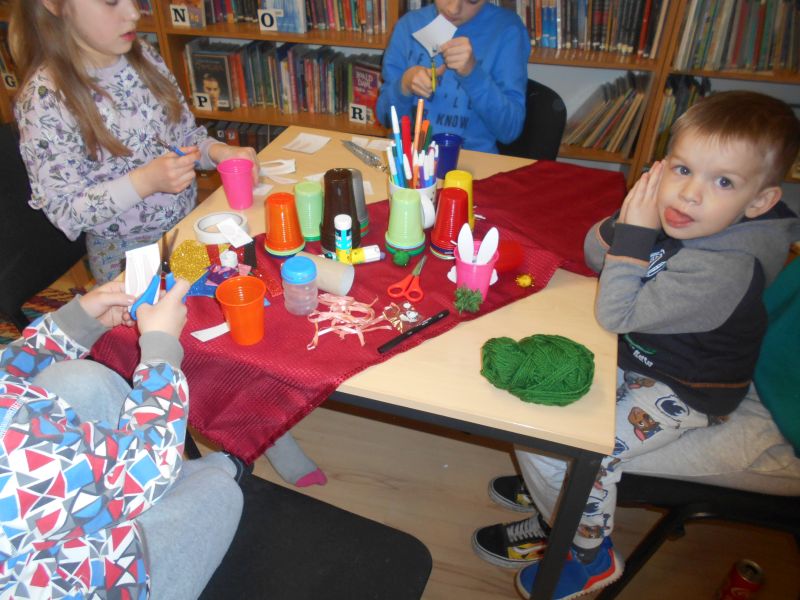 Uczestnicy zajęć siedząc przy stole wykonują prace plastyczną - zajączki z kubka plastikowego