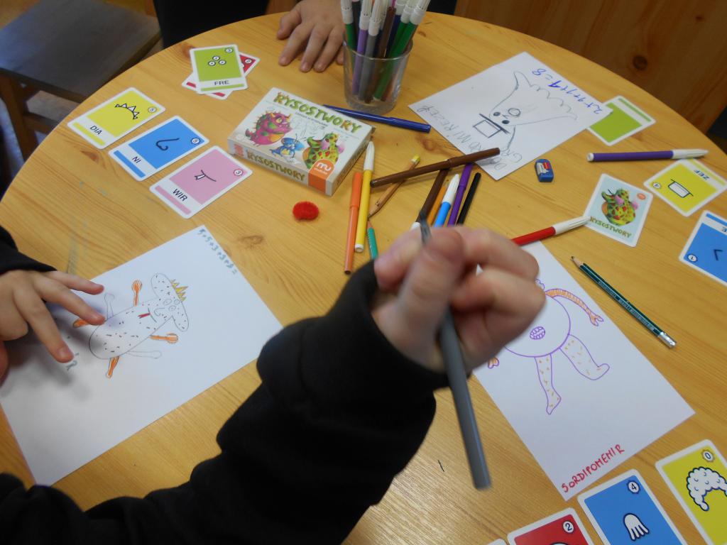 Dzieci w trakcie gry w Rysostwory; na stoliku leżą rozłożone karty, pisaki i kartki z narysowanymi stworkami 